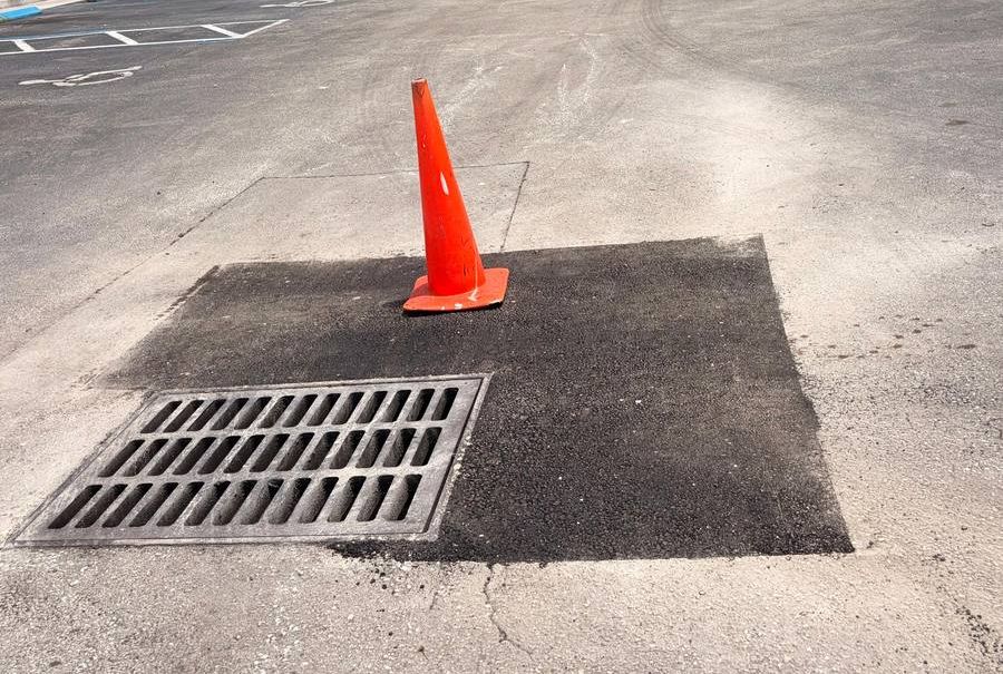Fresh asphalt patch around a storm drain grate in a commercial parking lot, marked with a cone